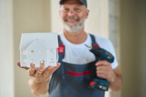 Photo of smiling construction worker in baseball cap and overalls holding house model in one hand and drill in other